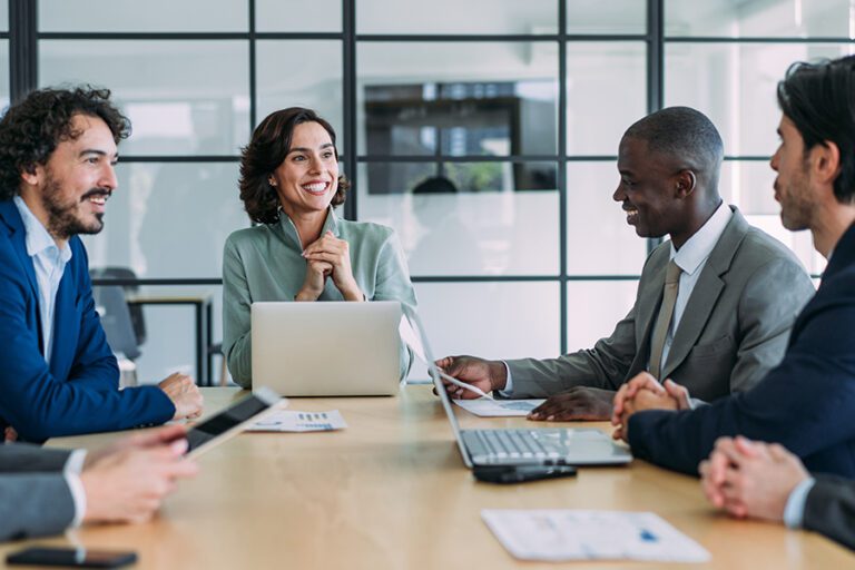 A group of five people in business attire sit at a conference table with laptops and documents, engaged in a meeting. One woman smiles while speaking to a man across from her.