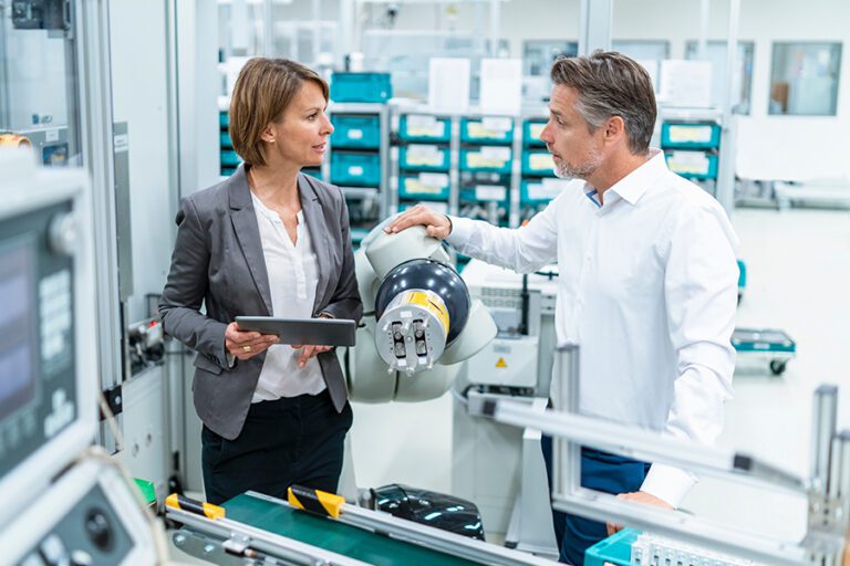 Two professionals stand in a modern factory. The woman holds a tablet, and the man rests his hand on a robotic arm, discussing the machinery.