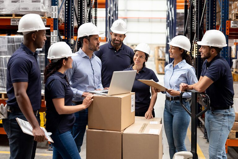 A group of workers wearing helmets and safety gear engage in a discussion around packages in a warehouse, with one person using a laptop and others holding clipboards.