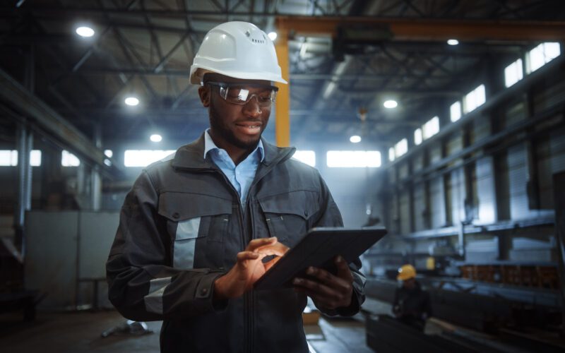 A man wearing a hard hat and safety glasses operates a tablet in an industrial warehouse setting.