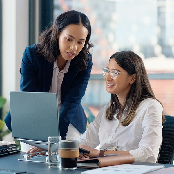 Two women in a modern office setting, one standing and the other sitting, both looking at a laptop screen. They appear to be collaborating on a project.