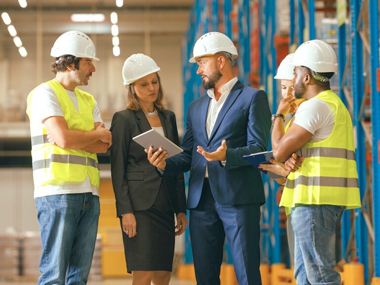 A group of five people, including three workers in safety vests and helmets, and two individuals in formal attire, stand discussing something in a warehouse. One person is holding a tablet.