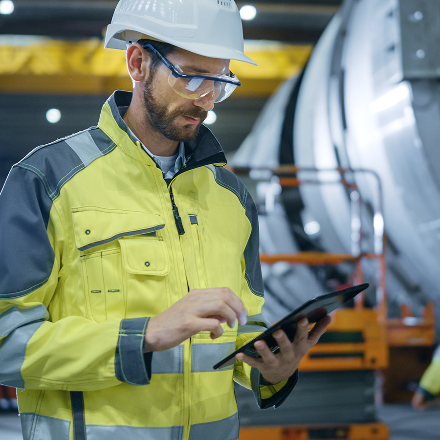 A man in a high-visibility jacket and hard hat operates a tablet in an industrial setting.