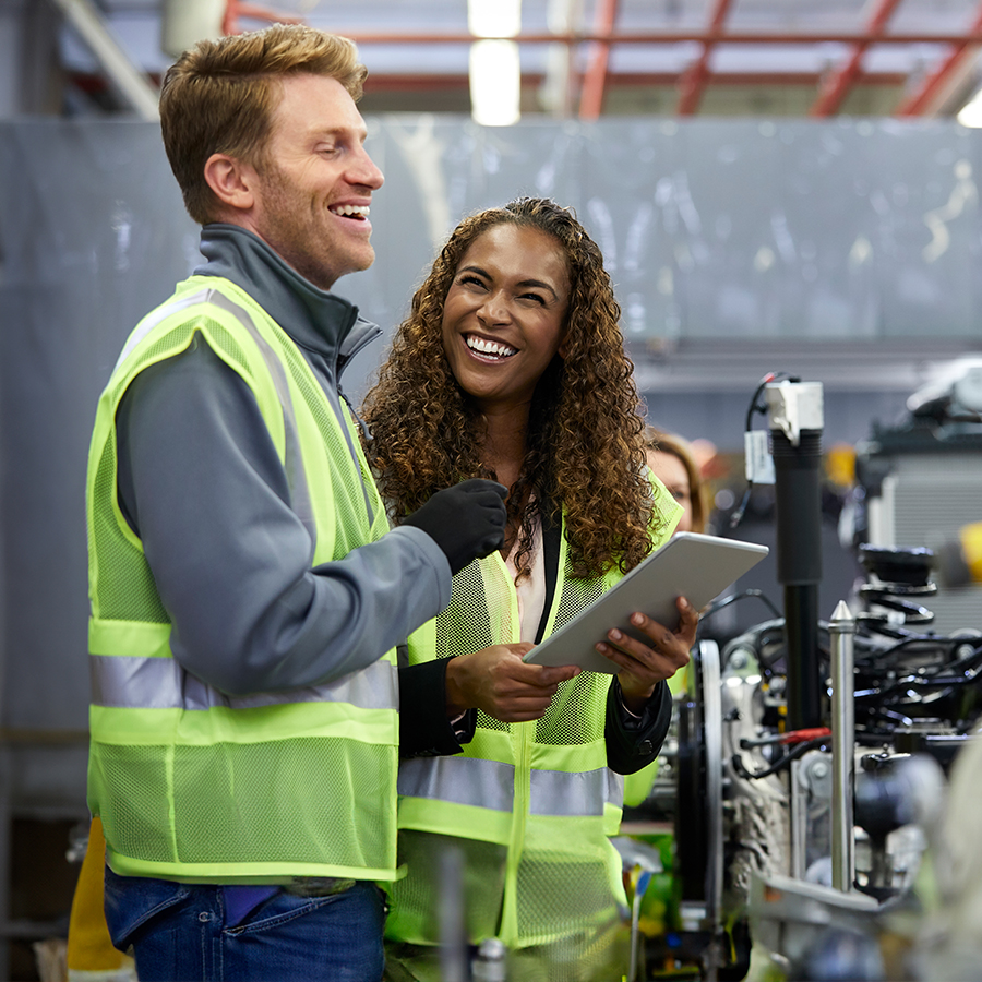 Two people wearing safety vests stand in an industrial setting, smiling and discussing something on a tablet.