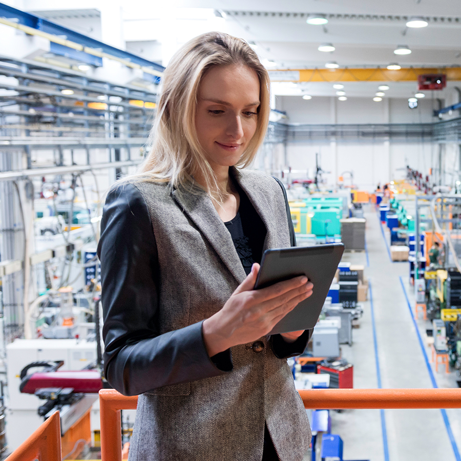 A woman stands in a factory, holding and looking at a tablet. She is wearing a grey blazer with black sleeves, and various machinery is visible in the background.