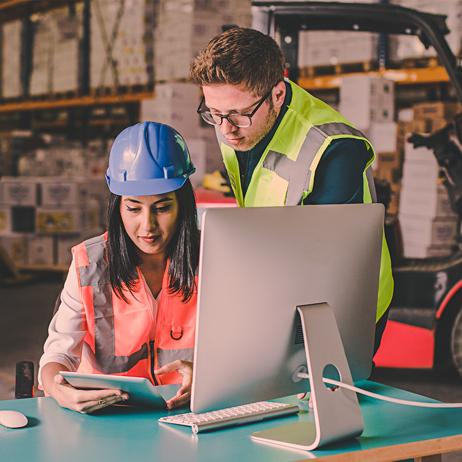 Two warehouse workers wearing safety vests and helmets are working together at a desk with a computer and tablet. They are inside a warehouse with shelves and a forklift in the background.