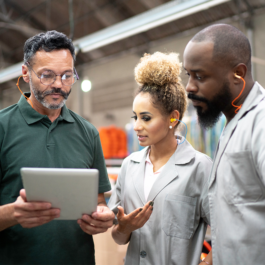 Three factory workers wearing ear protection look at a tablet while discussing something in a warehouse.