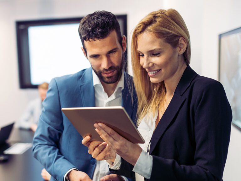 Two business professionals, a man and a woman, reviewing content on a tablet in an office setting. Both are dressed in formal attire, with the woman smiling as she holds the tablet.