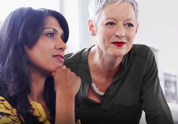 Two women engaged in a discussion, one with long dark hair wearing a yellow patterned top, the other with short gray hair wearing a green shirt. They appear to be in a bright, well-lit indoor setting.