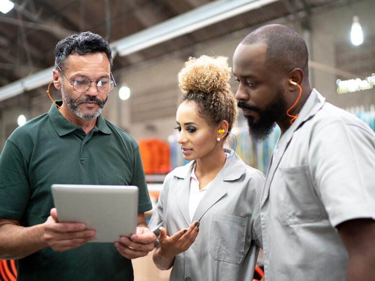 Three factory workers wearing ear protection discuss something on a tablet in an industrial setting.