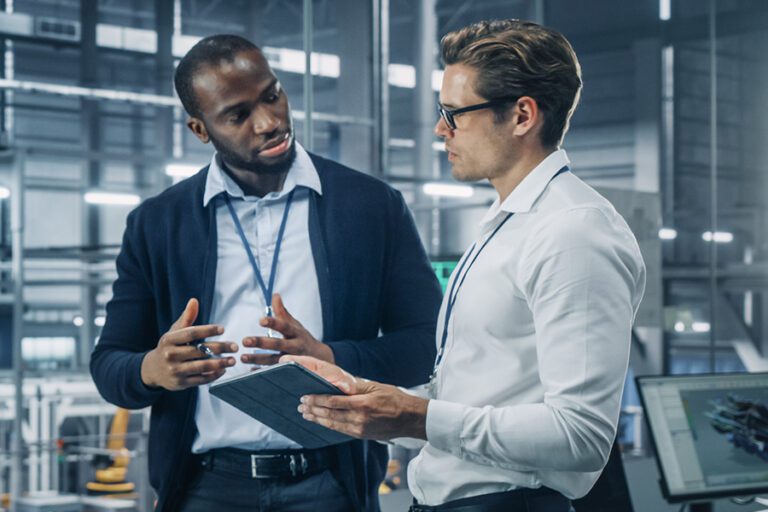 Two men in a modern office discuss workforce analysis software while holding a notebook and a pen. They are wearing ID badges and standing near a computer.