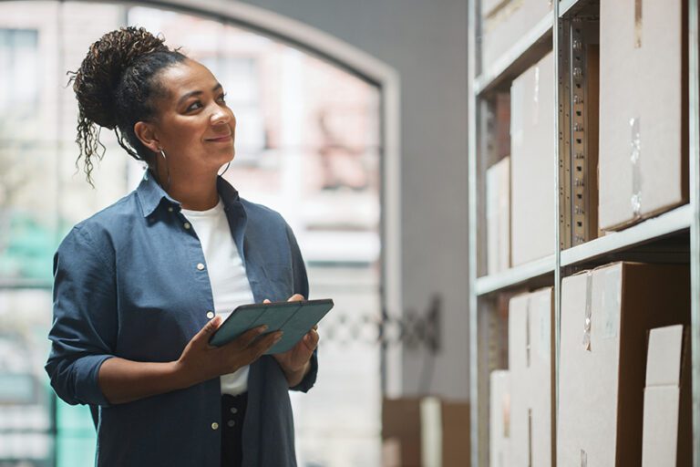 A person holding a tablet, equipped with workforce analysis software, looks at boxes on shelves in a well-lit room.