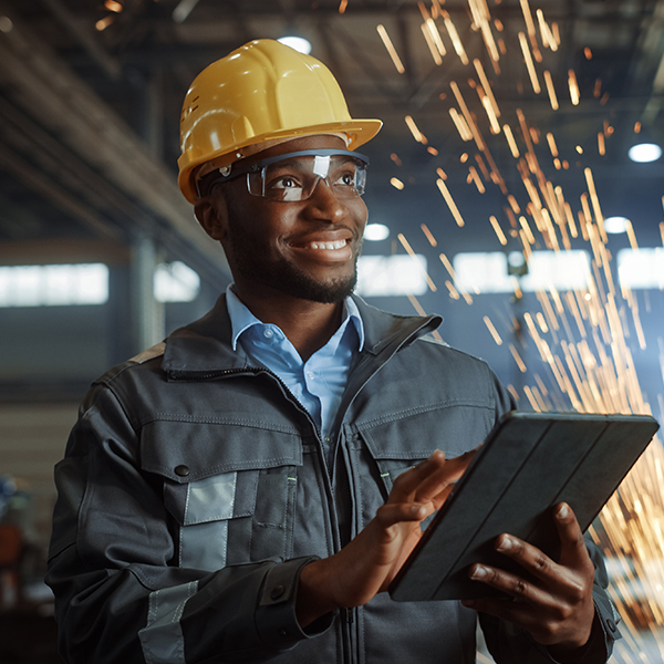 A worker in safety gear holds a tablet equipped with workforce analysis software, with sparks flying in the background inside an industrial setting.