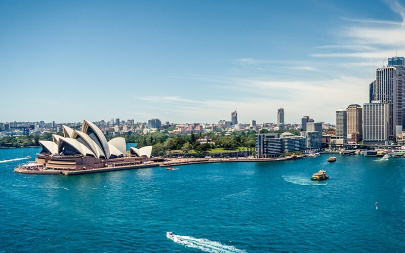 Aerial view of Sydney Harbour with the Sydney Opera House, ferries, and city skyline under a clear blue sky.