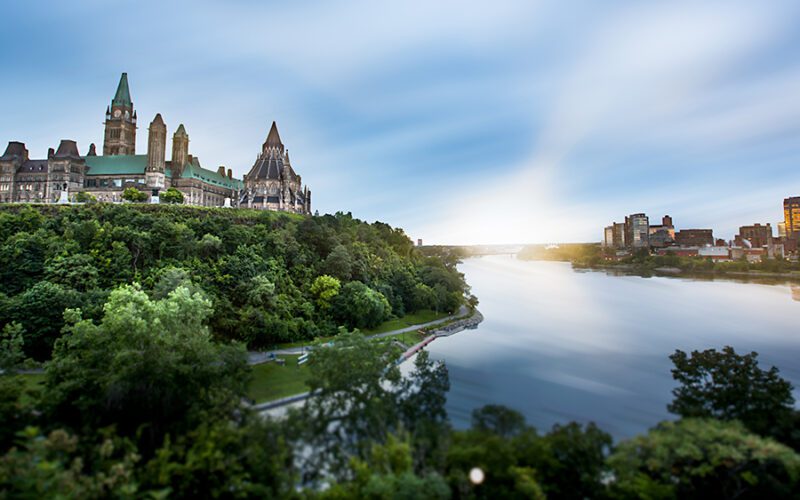 A scenic view of gothic-style buildings on a hill overlooking a river, with a city skyline in the distance under a bright sky.