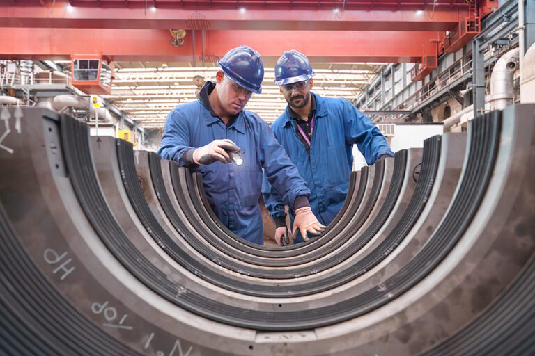 Two workers in blue uniforms and helmets inspect large metal rings in an industrial setting.