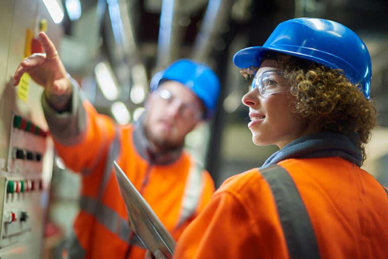 Two workers in orange safety vests and blue helmets operate control panels in an industrial setting.