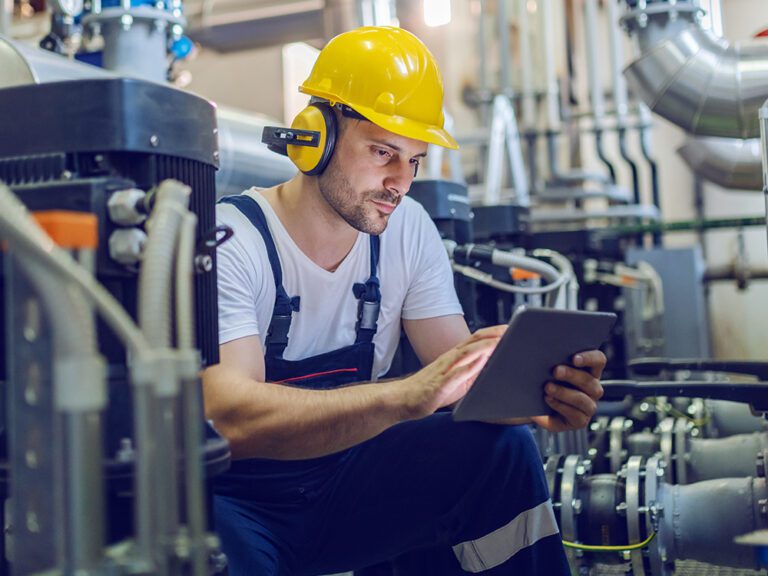 A worker in a hard hat and earmuffs uses a tablet while surrounded by industrial equipment and pipes.