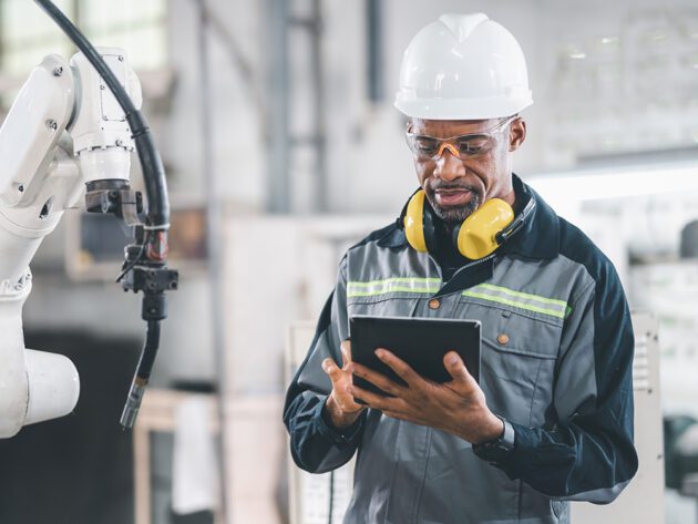 A worker in a hard hat and safety gear uses a tablet in an industrial setting, near robotic machinery.