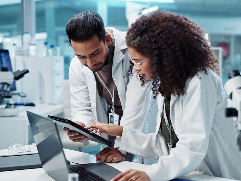 Two scientists in lab coats discuss data on a tablet and laptop in a laboratory setting.