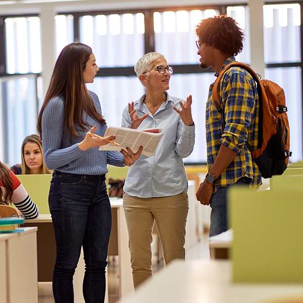 Three people are standing and talking in a classroom. One person is holding a book. Other students are seated in the background.