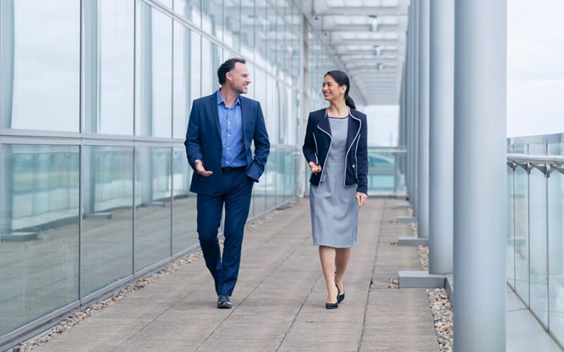 Two people in business attire walking and talking in a modern glass corridor.