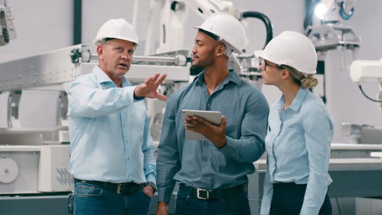 Three people in hard hats and blue shirts stand in a factory, engaged in discussion. One holds a tablet. Machinery is visible in the background.