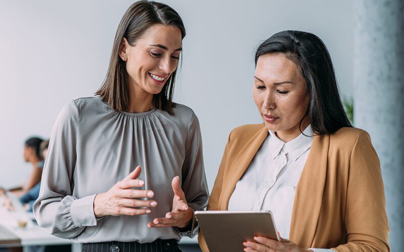 Two women in an office setting, one in a gray blouse gesturing while the other in a beige jacket holds and looks at a tablet.