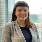 A person with glasses and a plaid blazer stands in front of a cityscape window.