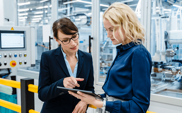 Two women in business attire discussing something on a tablet in a modern industrial setting.