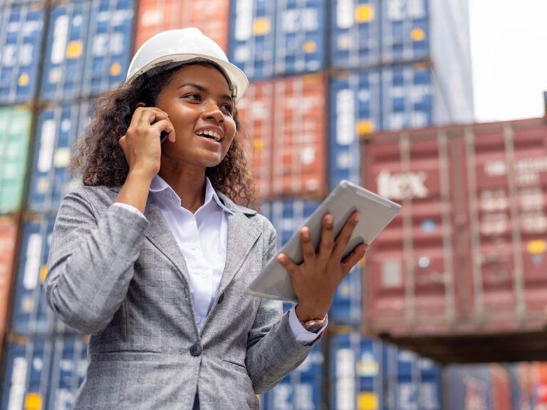 A woman in a hard hat talks on the phone while holding a tablet in front of stacked shipping containers.