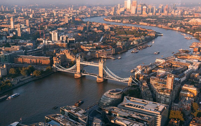 Aerial view of Tower Bridge over the River Thames in London, surrounded by urban buildings and boats on the water, during sunset.