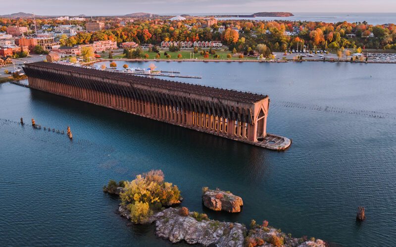 Aerial view of a long, wooden ore dock extending into a lake, surrounded by colorful autumn trees and urban buildings.