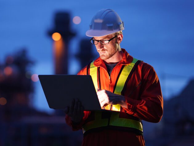 A worker in a hard hat and reflective vest uses a laptop at an industrial site during twilight.