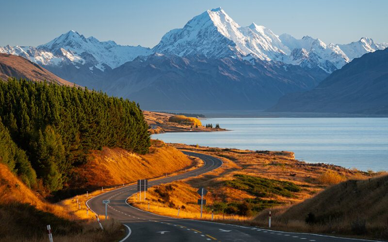 A winding road leads to snow-capped mountains, bordered by a lake on one side and dense pine forest on the other under a clear blue sky.