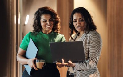 Two women stand together, looking at a laptop. One is holding a folder and a coffee cup.