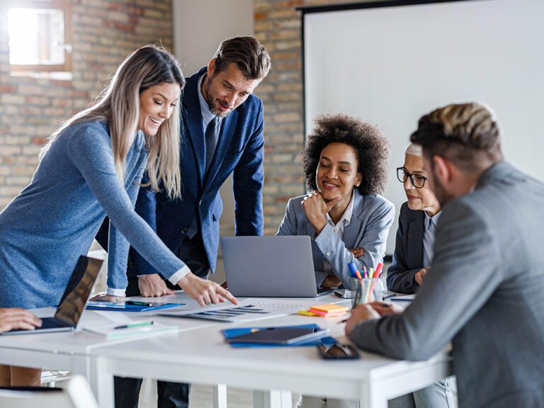 A group of professionals in a meeting, with one person pointing at documents on a table. Laptops and stationery are also on the table.