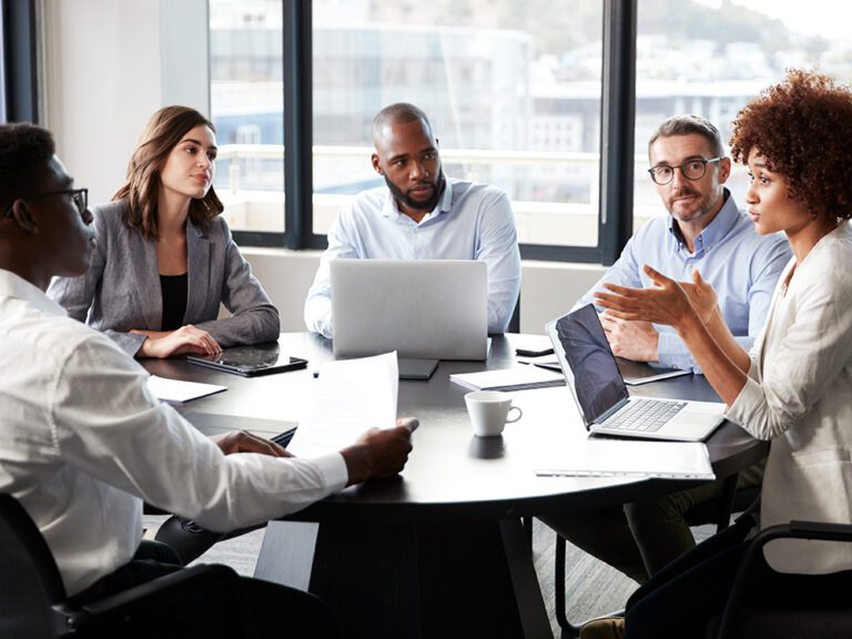 Five people in a meeting room sit around a table with laptops and documents, engaging in discussion.