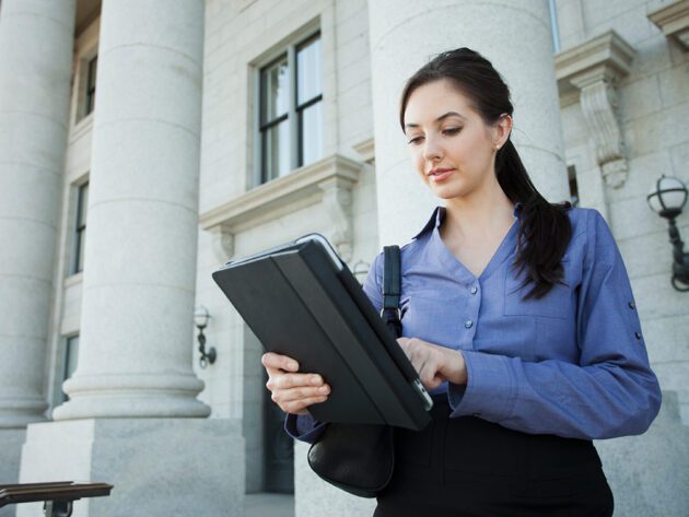 A woman stands outside a building with columns, looking at a tablet and holding a shoulder bag.