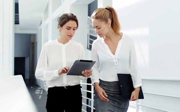 Two women in professional attire stand in a bright hallway. One holds a tablet while the other holds a folder.