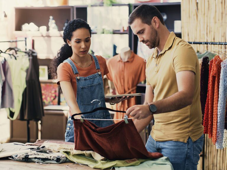 A man and woman browse clothing on hangers in a store. He holds a garment, while she appears to discuss or evaluate it. The store has racks of clothes and a casual, informal atmosphere.