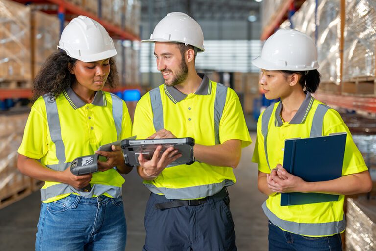 Three warehouse workers in high-visibility vests and hard hats discuss logistics using a handheld device and clipboard in an indoor storage area.