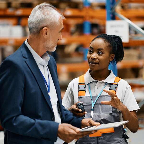 Two people in a warehouse setting, one in a suit holding a clipboard, the other in work overalls holding a device, engaged in conversation. Shelves with boxes are visible in the background.