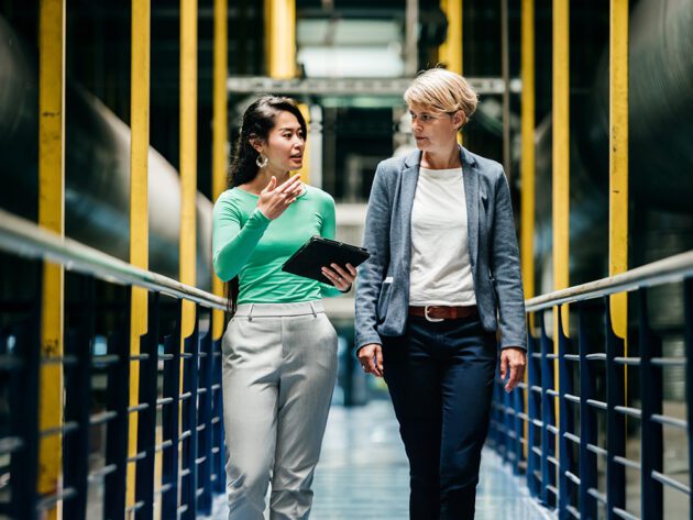 Two people walk through an industrial setting, discussing something. One holds a tablet. They are surrounded by yellow railings and large pipes.