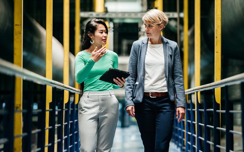Two people walk through an industrial setting, discussing something. One holds a tablet. They are surrounded by yellow railings and large pipes.