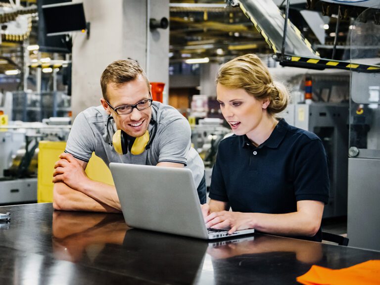 Two people in a workshop setting look at a laptop. The man leans on the table with ear protection around his neck. The woman types while sitting. Industrial equipment is in the background.