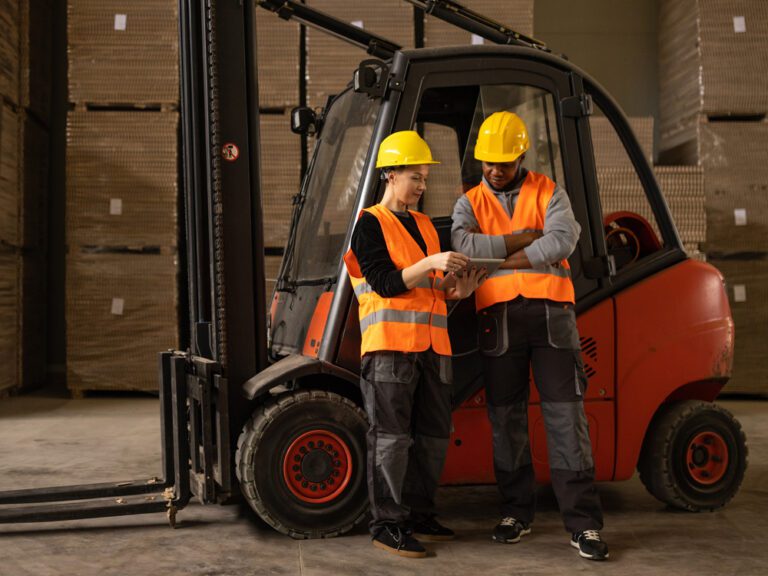 Two workers in orange safety vests and yellow helmets stand beside a forklift in a warehouse, examining a clipboard.