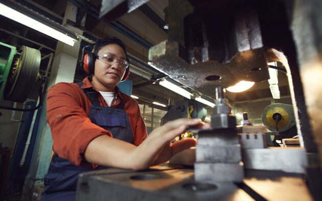 A person operates industrial machinery in a workshop, wearing safety gear including ear protection and goggles.