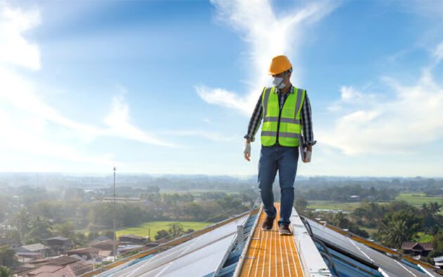 A construction worker wearing a safety vest and helmet walks on a rooftop under a clear blue sky.