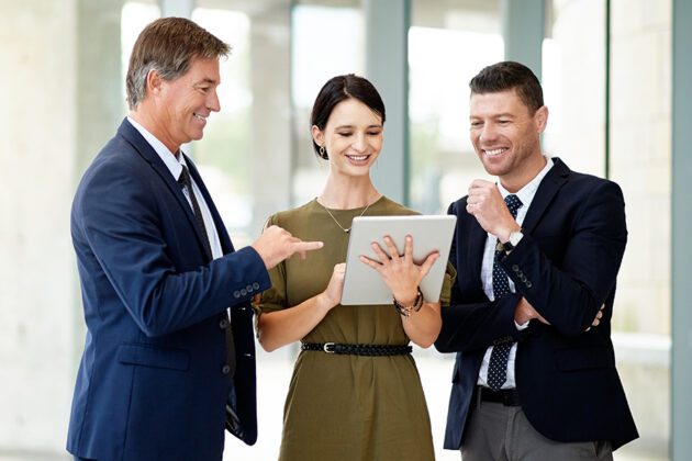 Three people in business attire smile and look at a tablet together in an office setting.